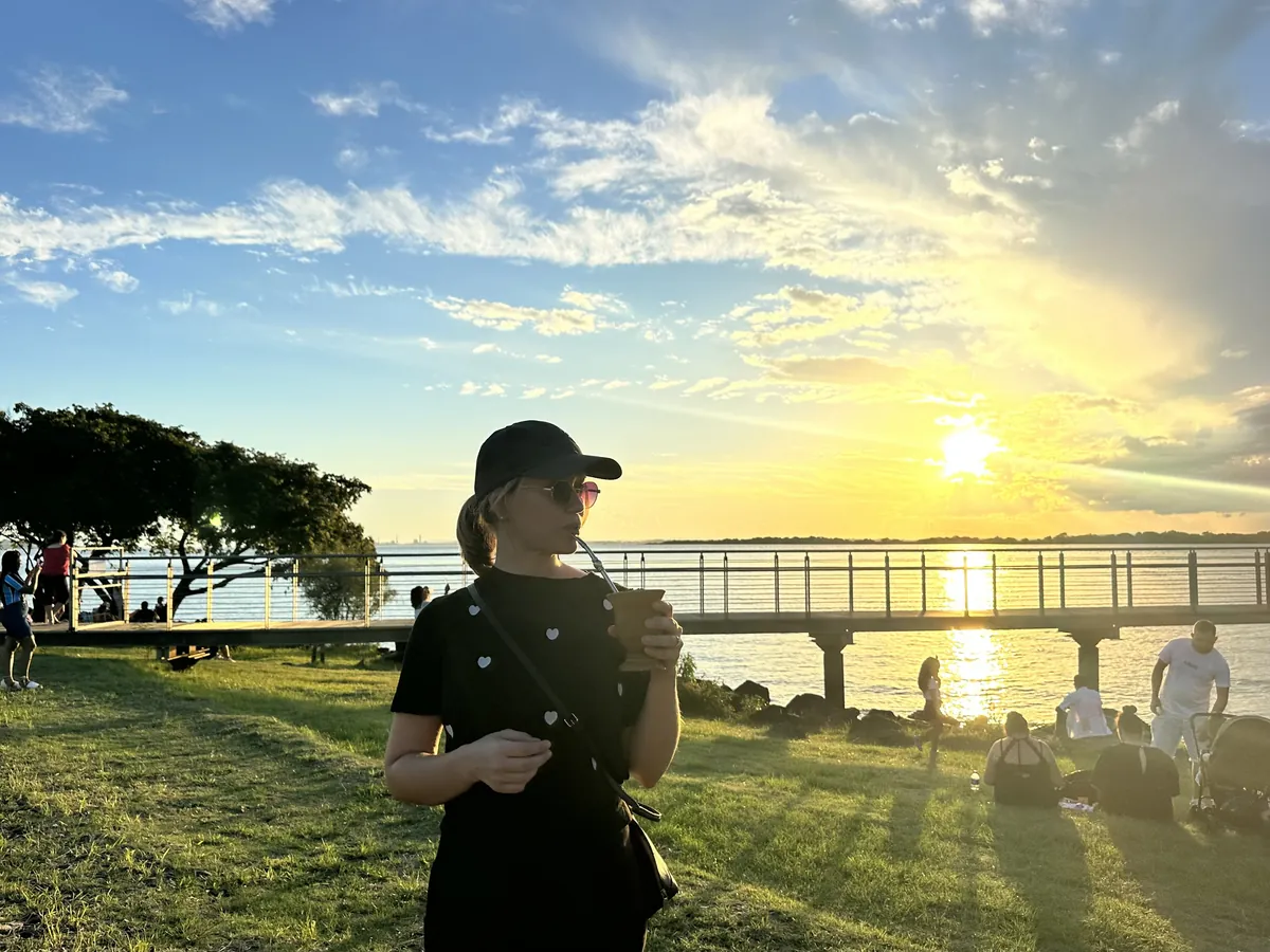 Ana drinking chimarr&atilde;o from a gourd at sunset on a grassy waterfront, a pier and the setting sun behind her