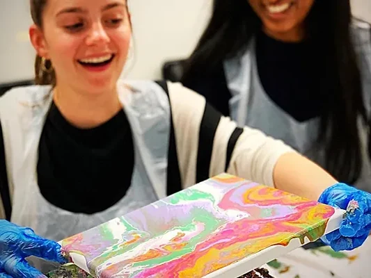 A woman laughing as she tilts her canvas and the poured paint runs in bright swirls